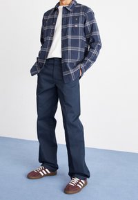 Camicia di flanella a quadri navy con bottoni dorati, t-shirt bianca a girocollo, pantaloni navy e sneakers marroni con strisce bianche.