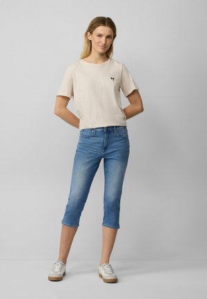 T-shirt beige a maniche corte con un piccolo logo nero, abbinato a jeans cropped blu chiaro e sneakers bianche con suola marrone.
