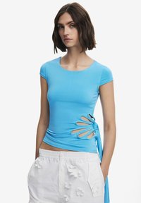 T-shirt ajusté bleu à col rond et manches courtes, doté d'ouvertures sur les côtés et d'un design froncé. Porté avec un short blanc texturé.