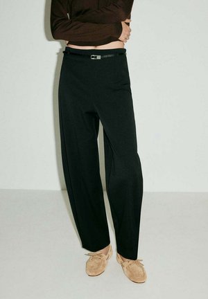 Persona che indossa pantaloni neri a vita alta con gambe larghe, una sottile cintura nera e scarpe beige in camoscio con lacci, in piedi davanti a uno sfondo neutro.