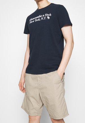 Henkilö, joka pukeutuu laivastonsiniseen Abercrombie & Fitch -t-paitaan ja beigeihinShortseihin, yhdellä kädellä taskussa, tasaisella valkoisella taustalla.