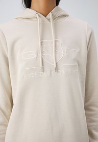 Sweat à capuche beige avec le logo en forme de bouclier "GANT 1949" et "American Sportswear" brodés sur le devant, porté par une personne.