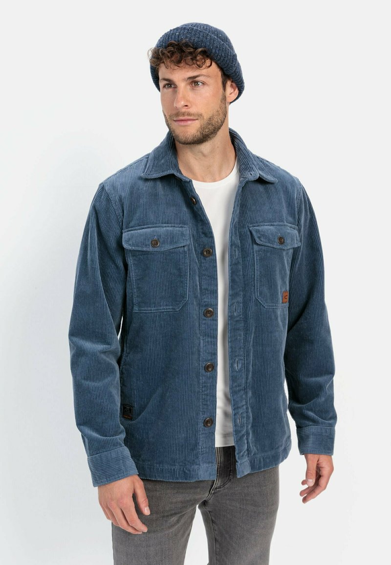 Blå cordjakke med frontknaplukning, to brystlommer og et tekstureret stof. Parret med en hvid skjorte og grå jeans.