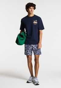 Camiseta de marina con texto y gráfico, pantalones cortos con estampado en azul y blanco, y una bolsa verde. El modelo lleva zapatillas deportivas grises.
