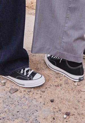 Schwarze Leinenschuhe mit weißen Gummisohlen und Schnürsenkeln. Ein Fuß ist mit dunkelblauen Jeans kombiniert, der andere mit hellgrauen Hosen.