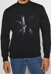 Felpa nera a maniche lunghe con logo lucido "CK Calvin Klein Jeans" sul petto, indossata con jeans in denim blu, su uno sfondo neutro.