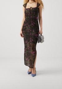 Schwarzes Blumenkleid mit kurzen Ärmeln, das eine knotbare Front, transparente Stoffe sowie lila und grüne Muster aufweist. Das Model hält eine graue Handtasche und trägt blaue High Heels.