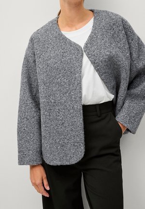 Femme portant un cardigan gris texturé sur une chemise blanche et un pantalon noir, avec une main dans la poche et l'autre détendue le long du corps.