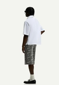 White short-sleeve shirt paired with gray shorts featuring a repeated pattern. Model wears black loafers and white socks.