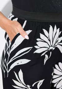 Černé kalhoty s výrazným bílým květinovým vzorem, mají elastický pás a boční kapsy. Hladká textura tkaniny, moderní design.