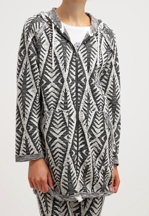 Person iført grå og sort lynlåshættejakke med geometrisk bladmønster, kombineret med matchende bukser og hvid skjorte under.