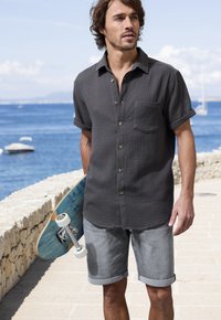 Kurzärmliges, dunkelgraues strukturiertes Hemd mit Knöpfen und einer Brusttasche, kombiniert mit hochgerollten grauen Shorts und einem blauen Skateboard.