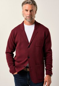 Burgunderfärgad stickad cardigan med sjalkrage, en bröstficka och två knappar, bärs över en vit skjorta och parats med blå jeans.