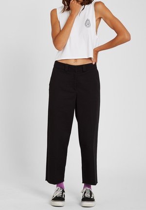 Person trägt ärmelloses weißes Crop-Top mit schwarzem Emblem, schwarze weite Hose, violette Socken und schwarze Sneakers und steht vor einem einfarbigen Hintergrund.