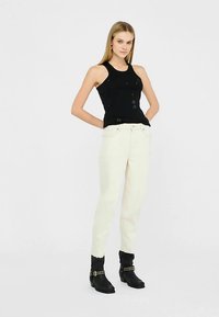 Top nero senza maniche con design strutturato, abbinato a jeans bianchi a vita alta e stivali neri al ginocchio con dettagli borchiati.