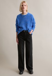 Blauer Pullover mit Textur und überschnittenen Schultern, kombiniert mit schwarzen, weit geschnittenen Hosen und klobigen schwarzen Schuhen. Einfache silberne Schichtungskette.