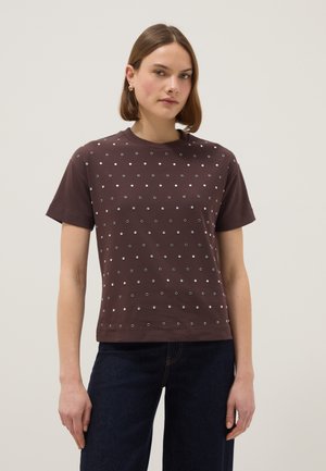 Femme portant un t-shirt marron à manches courtes avec de petits motifs d'œillets métalliques, associé à un jean bleu foncé sur un fond neutre.