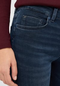 Sötétkék farmernadrág sima textúrával, klasszikus elülső zsebbel, kék gombbal és minimális varrási részletekkel.