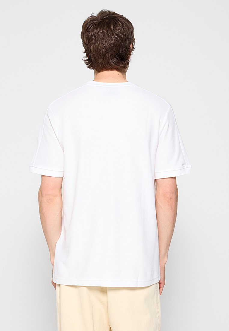 Mann mit braunen Haaren, der ein schlichtes weißes Kurzarm-T-Shirt und beige Hosen trägt, steht mit dem Rücken zu einem weißen Hintergrund.