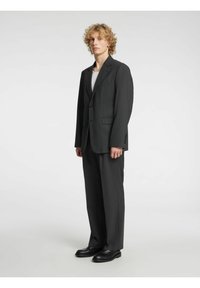 Mørkegrå dress med enbladsblazer, hakkede slag og rette bukser. Båret over en hvit skjorte, kombinert med svarte sko.