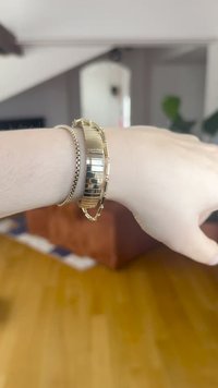 Aranyszínű fémszett óraszíj, szegmensekre tagolt kialakítással, polírozott és matt felületekkel, egy ezüst színű láncos karkötővel viselve a csuklón.