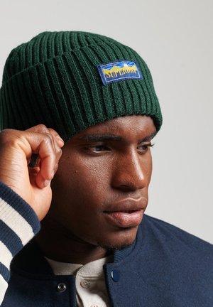 Beanie in maglia verde a coste con una toppa del logo "Superdry" blu e gialla, indossata da un modello. Lo sfondo è neutro e discreto.