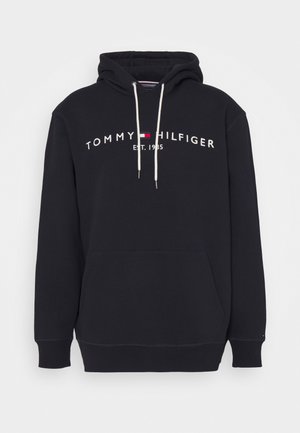 Schwarzer Kapuzenpullover mit weißen Kordeln, Fronttasche und dem Schriftzug "Tommy Hilfiger EST. 1985" mit rot-weiß-blauem Flaggenlogo auf der Brust.