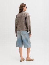 Persoon draagt een bruin suède jack, lichtblauwe denim shorts tot op de knie, beige veter schoenen en een donkere zonnebril, staand tegen een witte achtergrond.