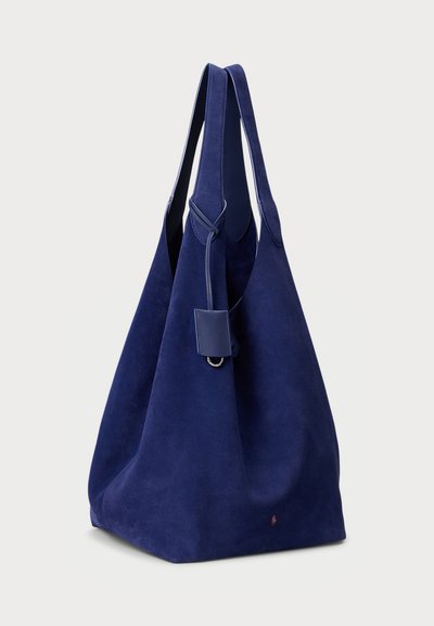 Sac fourre-tout en suède bleu marine avec une texture douce, de larges bretelles et un design épuré. Il est orné d'un accent en cuir et d'un détail de logo subtil près de la base.