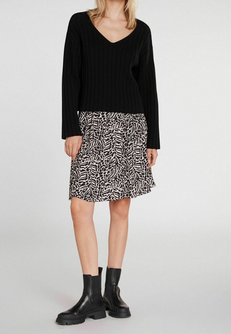 Femme portant un pull noir côtelé à col en V, une jupe imprimé animal longueur genoux et des bottines en cuir noires épaisses, debout sur un fond blanc.