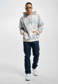 Sudadera gris claro con patrones geométricos azules, bolsillo frontal, cordones ajustables y puños beige. Combinada con jeans azul oscuro y zapatillas blancas.