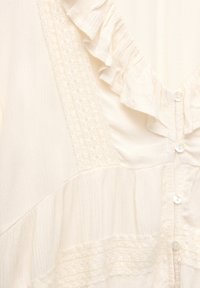 Blouse légère de couleur crème avec un décolleté en V, ornée de volants décoratifs, tissu texturé et fermeture boutonnée sur le devant.