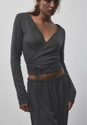 Le modèle porte un haut portefeuille gris anthracite à manches longues, noué à la taille, ainsi qu’un pantalon ample assorti, sur un fond de studio uni.