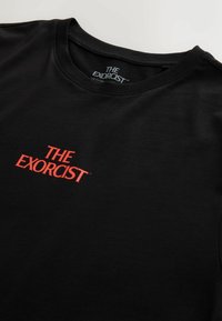 Melnā kokvilnas T-kreklā ir rakstīts "IZSITĒJS" treknajā sarkanā fontā, apaļa kakla dizains, gluda tekstūra un standarta izgriezums.