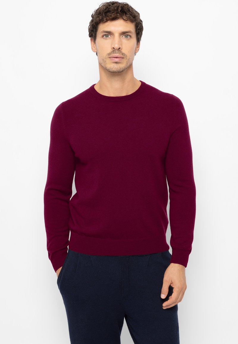 Bordeauxroter Rundhalspullover aus weichem Material mit langen Ärmeln und gerippten Bündchen. Wird über marineblauen Hosen getragen, keine sichtbaren Muster oder Logos.