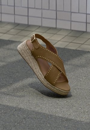 Sandale compensée en cuir marron avec semelle en jute tressé et clous dorés, posée sur l'asphalte près d'un mur carrelé.