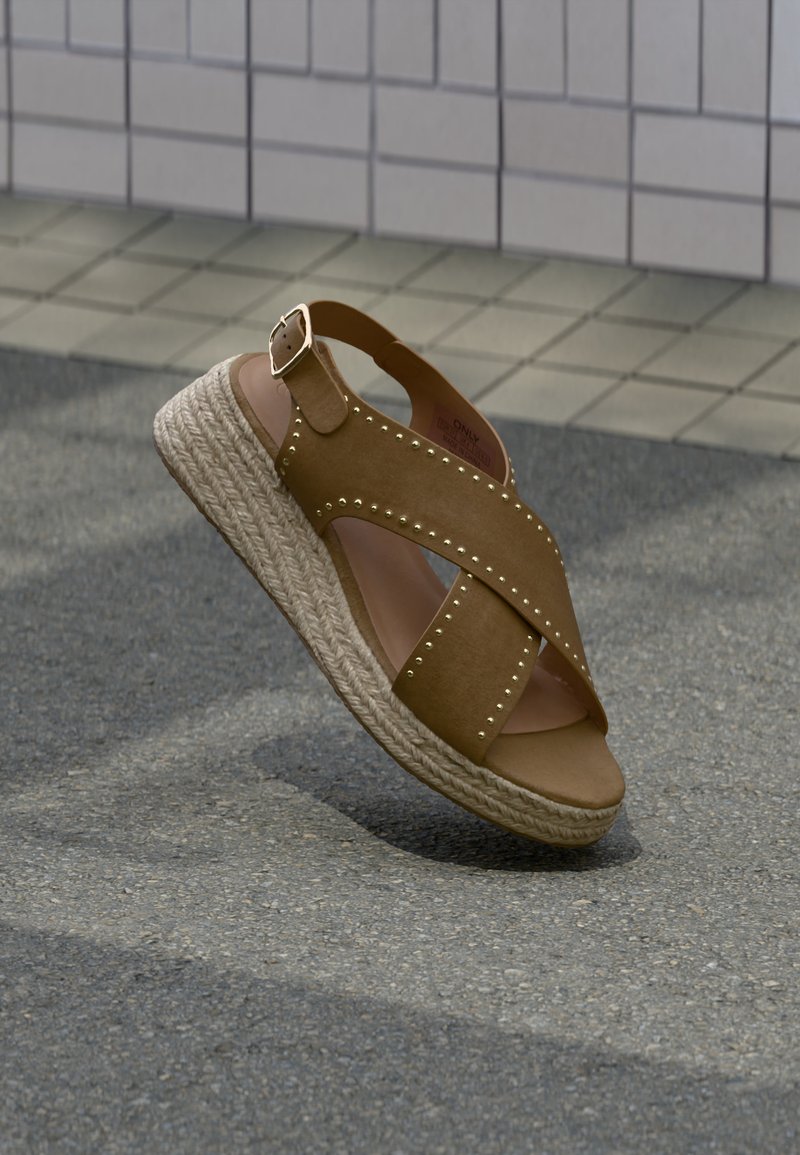 Καφέ δερμάτινο σανδάλι πλατφόρμα με πλεκτή σόλα από τζούτα και χρυσές τρουκς, τοποθετημένο σε άσφαλτο κοντά σε τοίχο με πλακίδια.