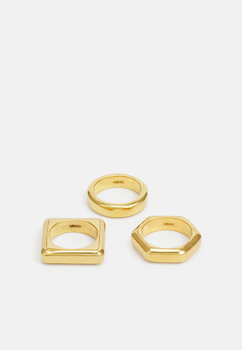 Trīs zelta gredzenu komplekts: viens apļa formā, viens kvadrātveida un viens sešstūra formā. Gluda virsma ar pulētu apdari, demonstrējot ģeometriskās formas.