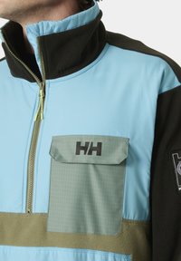 Fleece-Jacke mit einem hellblauen Oberteil, dunklen grünen Akzenten und einer strukturierten Brusttasche. Sie verfügt über einen Halb-Reißverschluss-Kragen und eine taschenklappe mit Karomuster.