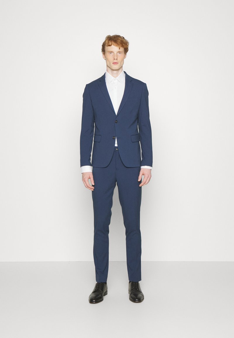 Completo blu navy su misura con vestibilità slim, giacca monopetto, revers a punta e camicia bianca. Tessuto strutturato, scarpe scure.