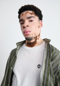 Grøn- og hvidstribet skjorte over en hvid t-shirt med et lille sort logo. Glat hud med tydelige mønstre. Kort, krøllet hår.