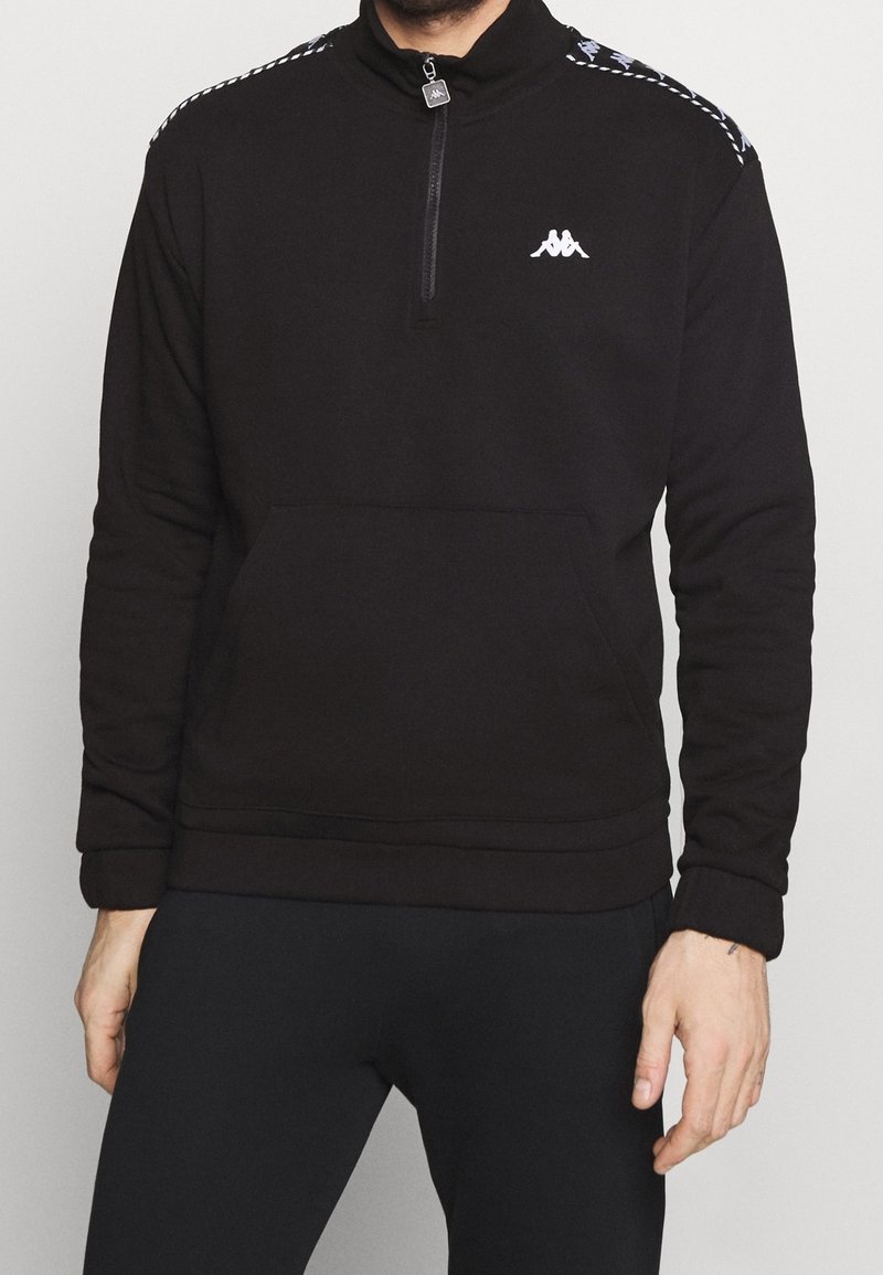 Homme portant un sweat-shirt noir à fermeture éclair quart de zip avec un logo blanc sur la poitrine et des bandes à motifs sur les épaules, assorti à un pantalon noir.
