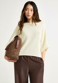 Strikket pullover i off-white, afslappet pasform med brede ærmer; parret med brune, lette, tætsiddende bukser; holder en vævet brun taske.
