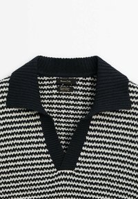 Pull en tricot à motif pied-de-poule noir et blanc avec un large col côtelé noir et une ouverture en V profonde, étiquette visible à l'intérieur du col.