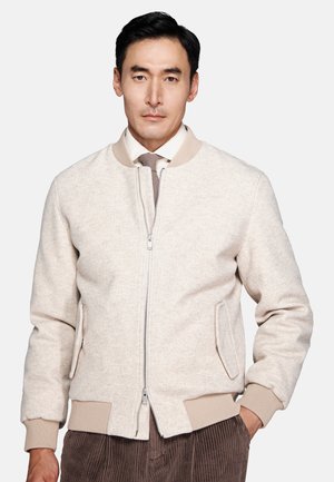 Man wearing a beige zip-up bomber jacket over a white shirt and tie, with brown corduroy pants, standing against a plain background.