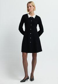 Vestito nero a pieghe con maniche lunghe, colletto bianco, chiusura a bottoni e dettagli argentati. Indossato con tacchi neri e collant trasparenti. Sfondo semplice.