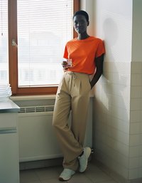 Oranžové tričko s krátkým rukávem, béžové kalhoty s vysokým pasem a bílé tenisky. Stojící u okna s žaluziemi, držící sklenici vody.