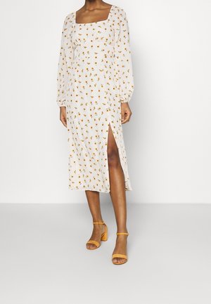 Femme portant une robe midi blanche à manches longues avec de petits imprimés de fleurs orange et une fente latérale, associée à des talons blocs jaunes ouverts à l'avant.