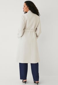 Lysegrå frakke med bælte i taljen, lange ærmer og en struktureret design. Glat stof med en medium længde, båret over mørke denim.