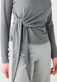 Haut gris à manches longues avec un design torsadé à l'avant, en tissu doux. Associé à un pantalon gris clair, présentant une texture lisse.